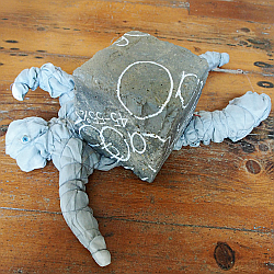 Ein Schlackestein, auf welchem die Chemikalien geschrieben sind. Unter dem Stein liegt eine Stoffpuppe eingequetscht